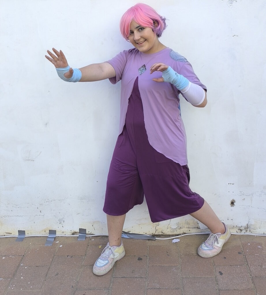 A girl dressed in a glimmer cosplay with a pink and purple wig, light purple shirt, sparkly blue cape, purple pants, and blue fingerless gloves.