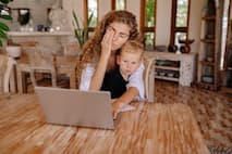 A mother using a laptop at a table with a child on her lap, visibly stressed.