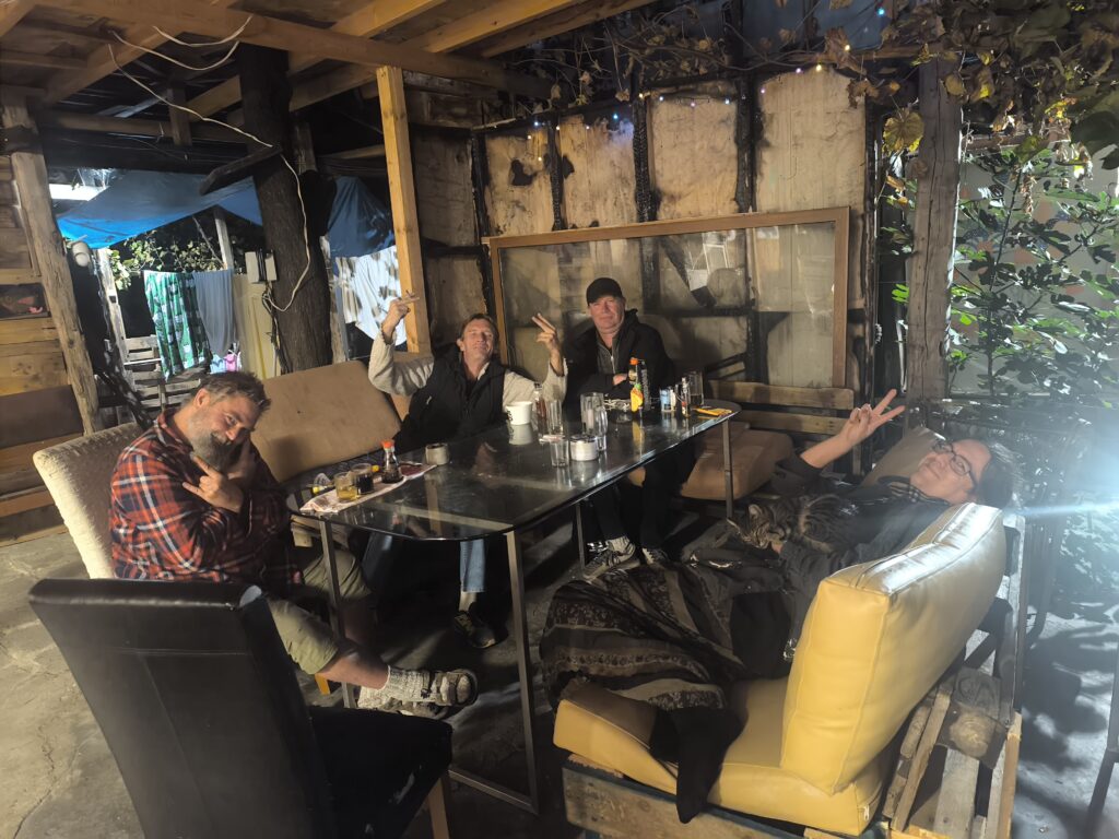 Four people sitting around a glass table in a rustic outdoor hostel area at night, with fairy lights and wooden beams. From left to right: Paul, James, another guest, and Jenny relaxing with a cat on her lap.