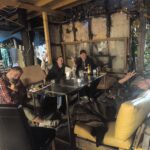Four people sitting around a glass table in a rustic outdoor hostel area at night, with fairy lights and wooden beams. From left to right: Paul, James, another guest, and Jenny relaxing with a cat on her lap.