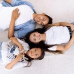A smiling family lying on their backs on a white textile surface, seen from above.